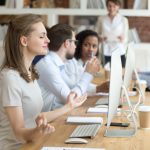 Peaceful millennial woman practice yoga in front of computer at workplace, calm female employee meditating controlling emotions and managing stress, worker sit in lotus position in office