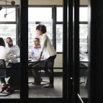 Millennial businesswoman stands addressing colleagues and thinking new strategy at a meeting, seen through glass wall
