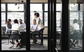 Millennial businesswoman stands addressing colleagues and thinking new strategy at a meeting, seen through glass wall