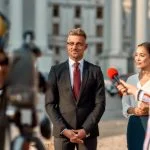 Horizontal shot of politician giving interview during campaign to journalists. People making interview using digital camera and equipment set at outdoor location.