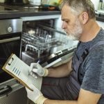 Male Technician Sitting Near Dishwasher Writing On Clipboard In Kitchen