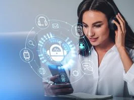 Businesswoman in headphones is holding smartphone with digital interface with various holograms having online conference call. Robot, smart watch, pie diagram. Office workplace with laptop, notebook
