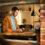 Handsome Man Typing on Laptop Computer in Loft Apartment, Sunny Day Outside. Creative Male Checking Social Media, Browsing Internet. Productivity in Working From Home.