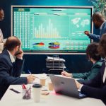 Startup team analyzing worldwide sales, shares and client feedback, using interactive boar to examine revenue and funds.