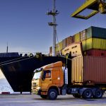Truck transportation carrying forty-foot container leaving port terminal with ship and quay crane on the background